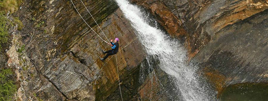 Homme avec casque et équipement de sécurité escaladant une paroi rocheuse avec une cascade en arrière plan.